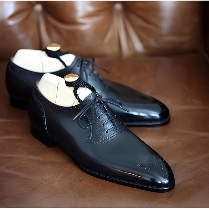 Pu&ograve; includere: Un paio di scarpe eleganti in pelle nera con finitura lucida, caratterizzate da un classico design stringato e perforazioni decorative. Le scarpe sono esposte su una superficie in pelle marrone, con forme di legno all'interno.