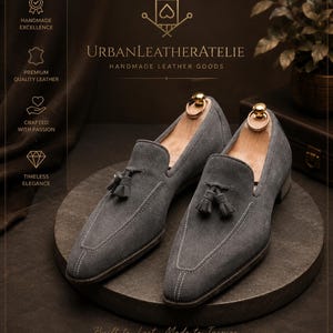 Może przedstawiać: Para szarych zamszowych mokasyn&oacute;w z chwostami, szpiczastym noskiem, białymi kontrastowymi szwami i włożonymi drewnianymi prawidłami. Tekst UrbanLeatherAtelie Handmade Leather Goods znajduje się nad butami wraz z ikonami.