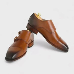 Könnte beinhalten: Ein Paar braune Leder-Doppelmonk-Schuhe. Die Schuhe haben ein Cap-Toe-Design und goldfarbene Schnallen. Das Leder hat einen Farbverlauf, der von einem helleren Braun zu einem dunkleren Braun an der Spitze übergeht.