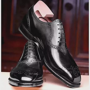 Può includere: Un paio di scarpe eleganti in pelle nera lucida con dettagli brogue e un classico design stringato. Le scarpe hanno una finitura lucida e sono esposte con una forma per scarpe in legno.