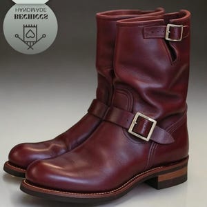 Puede incluir: Un par de botas de cuero burdeos con hebillas plateadas. Las botas son de media caña y tienen la punta redondeada. Las suelas son marrones con una costura de color claro alrededor del borde.