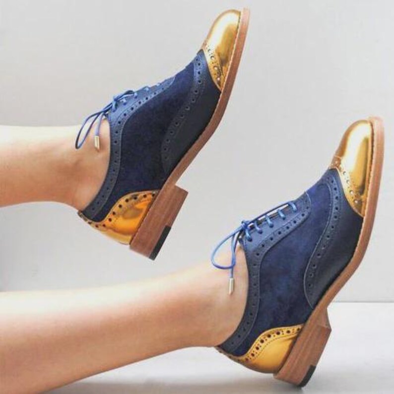 K&ouml;nnte beinhalten: Ein Paar marineblaue und goldfarbene Oxford-Schuhe mit Fl&uuml;gelspitzendesign. Die Schuhe haben goldfarbene Metallkappen und Fersenakzente, marineblaue Wildleder-Oberteile und blaue Schn&uuml;rsenkel. Holzsohle.