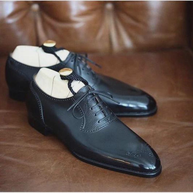 Pu&ograve; includere: Un paio di scarpe eleganti in pelle nera con finitura lucida. Le scarpe presentano un classico design stringato, decorazioni traforate e forme in legno. Le scarpe sono esposte su una superficie marrone.