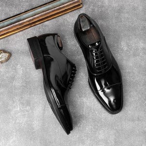 Puede incluir: Un par de zapatos de vestir negros de cuero brillante con puntera y cierre de cordones. Los zapatos se exhiben sobre una superficie gris, mostrando su acabado pulido y diseño clásico. Talla 43 (EU).