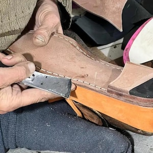May include: A shoemaker uses a blade to work on a leather shoe. The shoe has a tan upper, an orange sole, and a black toe cap. Other shoe components are visible in the background.