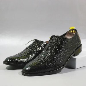 May include: A pair of black leather dress shoes with a textured, crocodile-like pattern. The shoes have a classic lace-up design and a glossy finish. The shoes are displayed on a white surface.