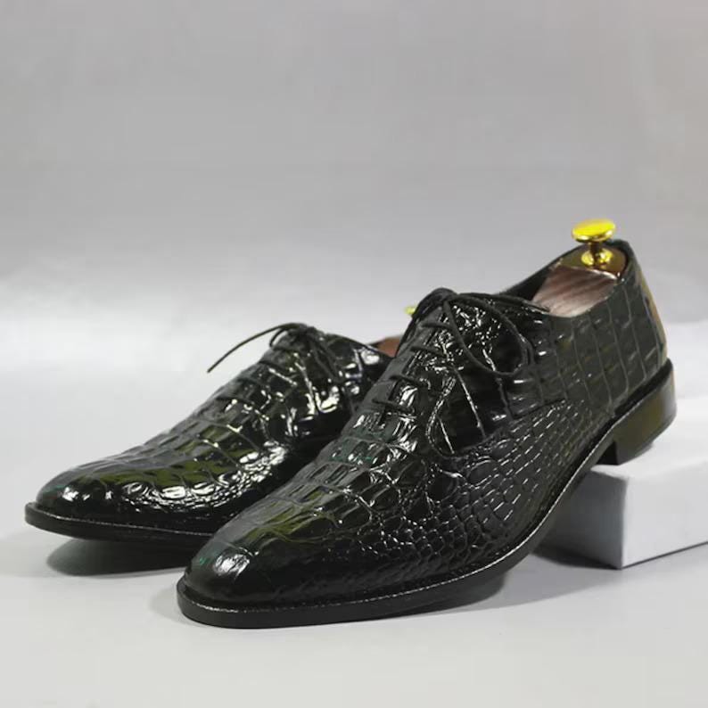 May include: A pair of black leather dress shoes with a textured, crocodile-like pattern. The shoes have a classic lace-up design and a glossy finish. The shoes are displayed on a white surface.