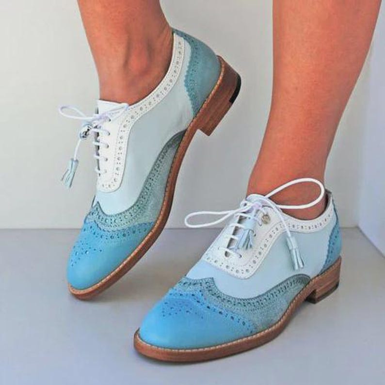 K&ouml;nnte beinhalten: Hellblaue und wei&szlig;e Oxford-Schuhe aus Leder mit Lochmuster. Die Schuhe haben ein klassisches Schn&uuml;rdesign mit Quasten und eine Holzsohle. Das Farbschema ist eine Mischung aus Hellblau, Wei&szlig; und einem dunkleren Blau an der Spitze.