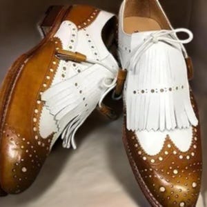 May include: A pair of two-tone leather dress shoes. The shoes feature a white and brown color scheme with decorative perforations and fringe detailing. The shoes have a classic, formal design.