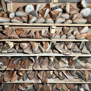 May include: A collection of wooden shoe lasts, in various sizes and shapes, neatly arranged on multiple shelves. The lasts are primarily brown with some showing signs of wear, suggesting age and use. The image conveys a vintage, workshop aesthetic.