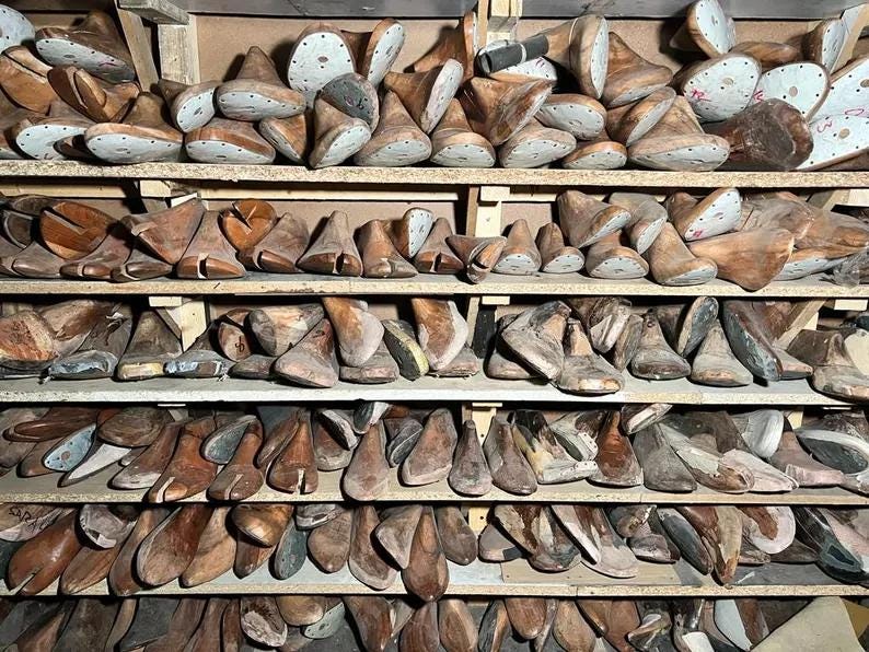 May include: A collection of wooden shoe lasts, in various sizes and shapes, neatly arranged on multiple shelves. The lasts are primarily brown with some showing signs of wear, suggesting age and use. The image conveys a vintage, workshop aesthetic.