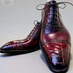 May include: A pair of burgundy leather dress shoes with a glossy finish. The shoes feature a textured, crocodile-like pattern on the toe and heel, with black laces. The shoes are designed with a classic, formal style.