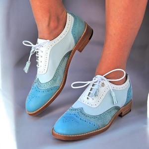 K&ouml;nnte beinhalten: Zweifarbige blaue und wei&szlig;e Oxford-Schuhe mit dekorativen Perforationen. Die Schuhe haben eine hellblaue Spitze und Ferse, eine wei&szlig;e Mitte und eine Holzsohle. Wei&szlig;e Schn&uuml;rsenkel und Quasten vervollst&auml;ndigen das Design.
