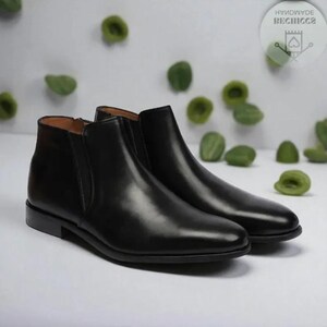 May include: A pair of black leather Chelsea boots. The boots have a sleek, polished appearance with a rounded toe and elastic side panels. The background features green accents and a white surface.