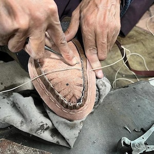 May include: A cobbler's hands working on a shoe sole. The leather sole is light brown with a darker brown trim. A needle and thread are used to stitch the sole. Tools and fabric are in the background.