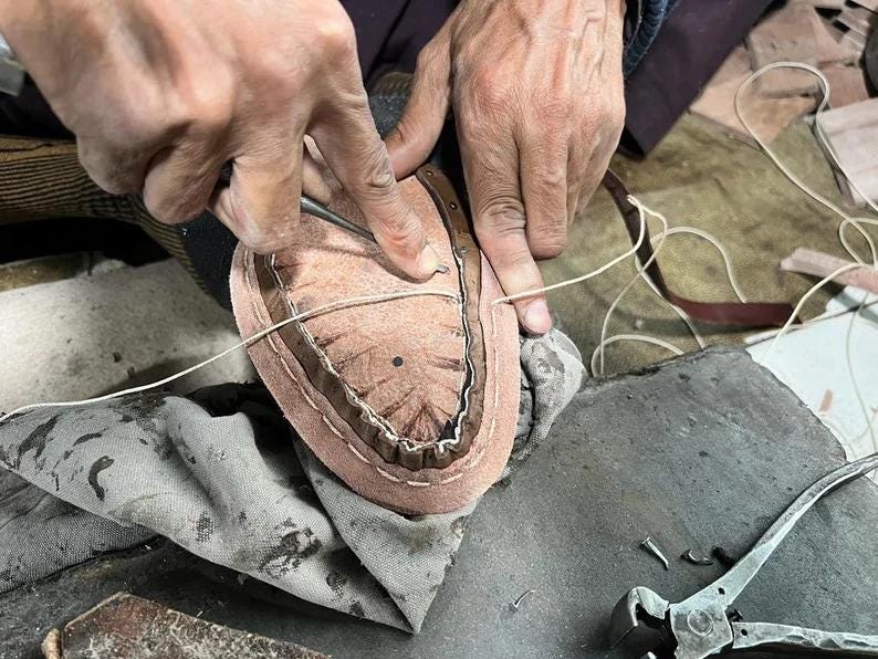 May include: A cobbler's hands working on a shoe sole. The leather sole is light brown with a darker brown trim. A needle and thread are used to stitch the sole. Tools and fabric are in the background.