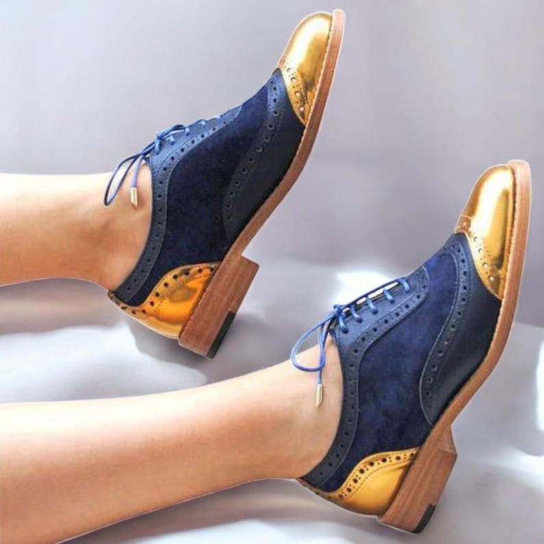 K&ouml;nnte beinhalten: Zweifarbige Oxford-Schuhe mit marineblauen Wildleder-Obermaterialien und goldenen metallischen Zehen- und Fersenakzenten. Die Schuhe haben blaue Schn&uuml;rsenkel und einen Holzabsatz. Stilvolle Schuhe.