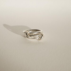 May include: A silver ring featuring a knot design. The ring is crafted from a shiny metal and has a simple, elegant style. The ring is set against a light-coloured background.
