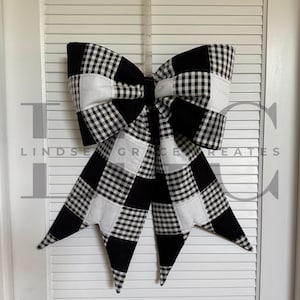 Peut inclure: Un grand nœud décoratif en tissu à carreaux noirs et blancs, suspendu à une porte blanche. Le nœud présente un motif patchwork avec un mélange de carrés blancs unis et à carreaux. Le nœud est suspendu par un cintre en plastique transparent.