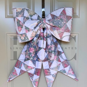 Pode incluir: Um grande laço decorativo feito de tecido acolchoado com um padrão de patchwork de triângulos brancos e estampas florais em tons de rosa, verde sálvia e azul. O laço tem duas alças largas e duas pontas longas. O texto LINDSEY CREATES é visível.