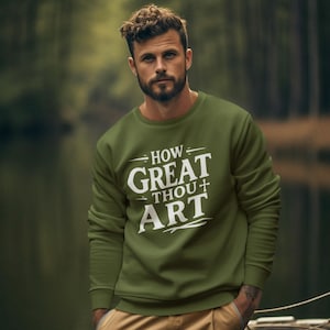 May include: Olive green sweatshirt with the text "HOW GREAT THOU ART" in white. The sweatshirt is long-sleeved and has a crew neck. The person is wearing khaki cargo pants. The background is a blurred outdoor scene.