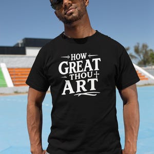 May include: Black t-shirt with the white text "HOW GREAT THOU ART". The shirt is a crew neck style and is being worn by a person wearing sunglasses. The background is a blue sky and a sports field.