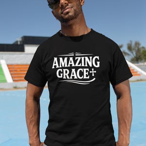 May include: Black t-shirt with the words "AMAZING GRACE +" in white, featuring a cross. The shirt is worn by a person wearing sunglasses. The background is a blue sky and a blue and white surface.