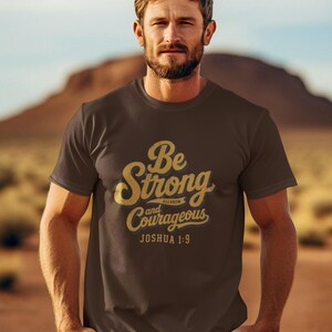 May include: A dark brown short-sleeved t-shirt featuring gold-colored text that reads Be Strong and Courageous JOSHUA 1:9. The shirt has a crew neckline and a standard fit, with the graphic printed prominently across the center of the chest area.