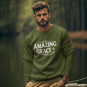 May include: Olive green sweatshirt with the text "AMAZING GRACE+" in white. The long-sleeved top is paired with khaki cargo pants. The image is set outdoors with a blurred background of trees and water.