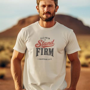 May include: A cream-colored short-sleeve t-shirt featuring the text "ALL4HIM" in small black letters, "Stand" in a large red distressed font, "FIRM" in a large black distressed font, and "1 CORINTHIANS 16:13" in small black letters.