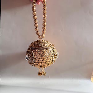 May include: A spherical gold-toned handbag featuring an intricate metallic mesh texture, embellished with clear crystals and a beaded handle. The bag has a hinged lid and a small dangling tassel at the bottom, showcasing detailed craftsmanship.