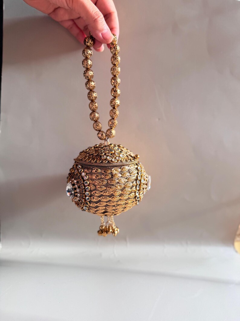May include: A spherical gold-toned handbag featuring an intricate metallic mesh texture, embellished with clear crystals and a beaded handle. The bag has a hinged lid and a small dangling tassel at the bottom, showcasing detailed craftsmanship.
