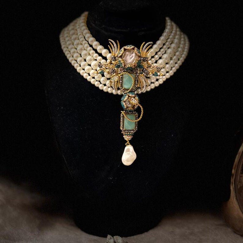 May include: A multi-strand pearl necklace with a decorative pendant. The pendant features gold filigree, green and clear gemstones, and a large baroque pearl drop. The necklace is displayed on a black mannequin.