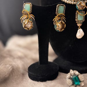 May include: Elegant jewelry set featuring gold-toned earrings with square, light green stones and pearl accents. The earrings are displayed on a black stand, alongside a matching necklace and ring. The set showcases intricate detailing and a luxurious aesthetic.