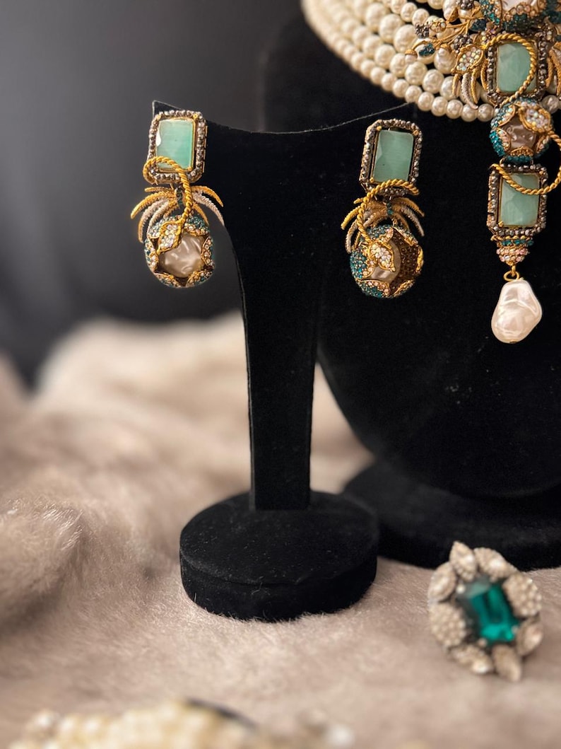 May include: Elegant jewelry set featuring gold-toned earrings with square, light green stones and pearl accents. The earrings are displayed on a black stand, alongside a matching necklace and ring. The set showcases intricate detailing and a luxurious aesthetic.