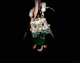 Puede incluir: Primer plano de un pendiente decorativo. El pendiente presenta un pequeño diseño de bolso adornado con detalles de cristal verde esmeralda y transparente. Una gran cuenta verde redonda cuelga debajo. Sobre un fondo oscuro.