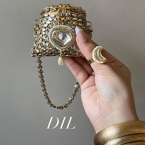 Op de afbeelding: Een goudkleurige, sierlijke handtas met een hartvormig middelpunt en een pareldruppel. Een halve maan ring met parelaccenten is ook zichtbaar. Het woord "DIL" is in witte letters onderaan de afbeelding gedrukt.