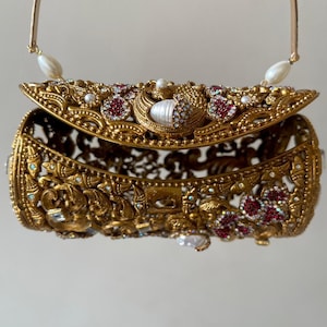 Puede incluir: Clutch dorada ornamentada con intrincadas tallas y adornos. La parte superior con bisagras está adornada con piedras preciosas rojas y transparentes, perlas y un diseño de pavo real. Un asa dorada con detalles de perlas completa el diseño. Mide unos 15 cm de largo.
