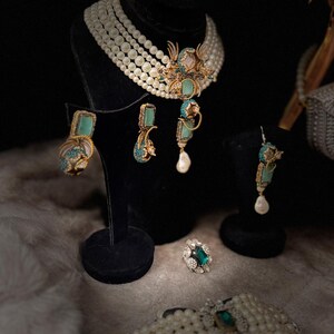 May include: An elegant jewelry set featuring a pearl necklace, earrings, a ring, and a bracelet. The necklace and bracelet have large pearls. The earrings and ring have turquoise and emerald accents. The jewelry is displayed on black stands.