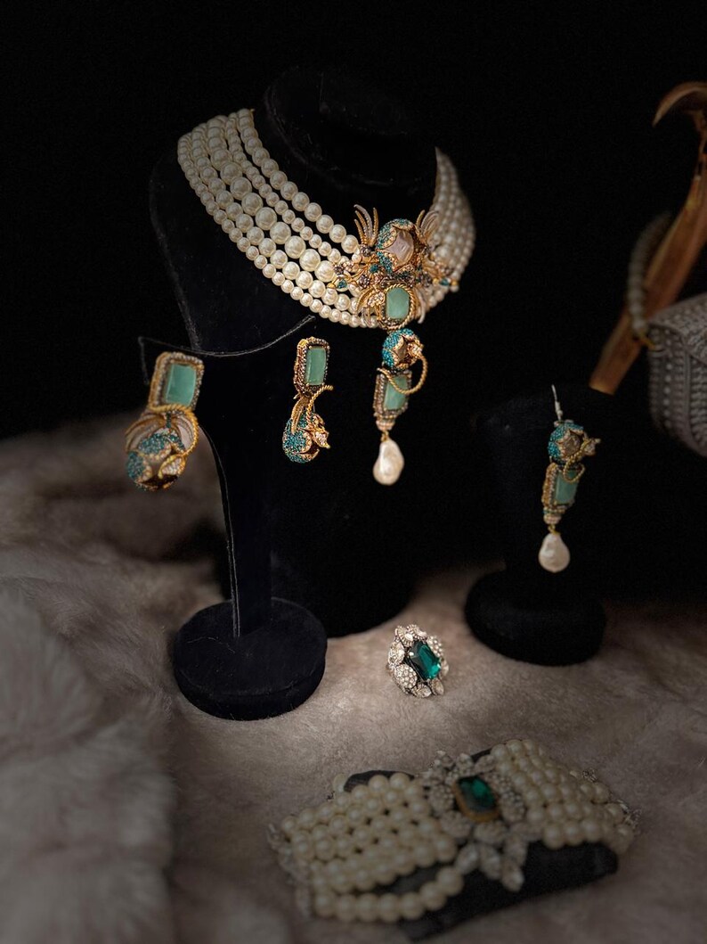 May include: An elegant jewelry set featuring a pearl necklace, earrings, a ring, and a bracelet. The necklace and bracelet have large pearls. The earrings and ring have turquoise and emerald accents. The jewelry is displayed on black stands.