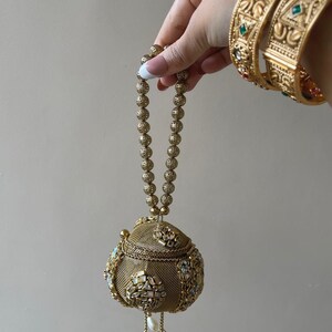 May include: A spherical gold-toned metal handbag featuring a fine mesh texture, ornate stone-encrusted floral motifs, and a beaded handle. The bag includes dangling pearl and metal charms at the base, complemented by matching wide gold-toned bangles.