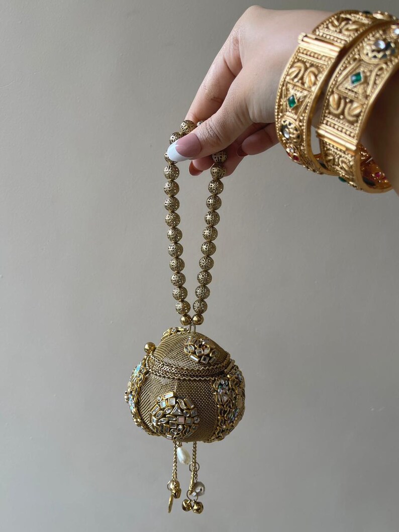 May include: A spherical gold-toned metal handbag featuring a fine mesh texture, ornate stone-encrusted floral motifs, and a beaded handle. The bag includes dangling pearl and metal charms at the base, complemented by matching wide gold-toned bangles.
