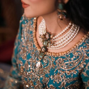 May include: A close-up of a teal and gold embroidered dress with a multi-strand pearl necklace. The necklace features a decorative pendant with green and gold accents. The dress has intricate gold embroidery and sequin details.