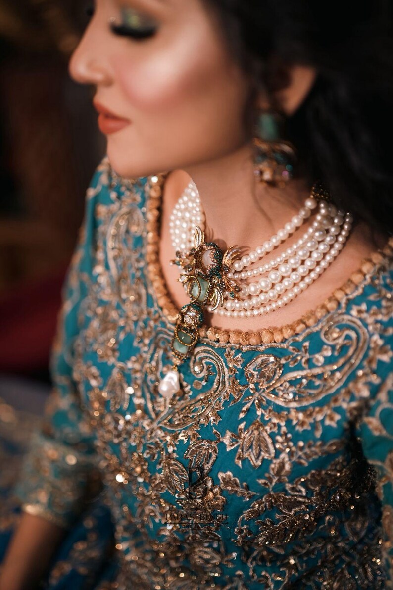 May include: A close-up of a teal and gold embroidered dress with a multi-strand pearl necklace. The necklace features a decorative pendant with green and gold accents. The dress has intricate gold embroidery and sequin details.