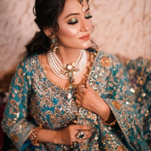 May include: A teal and gold embroidered ensemble, featuring a detailed bodice and sleeves. The outfit is adorned with gold embellishments and paired with a multi-strand pearl necklace, earrings, and a headpiece. A large ring with a green stone is visible.