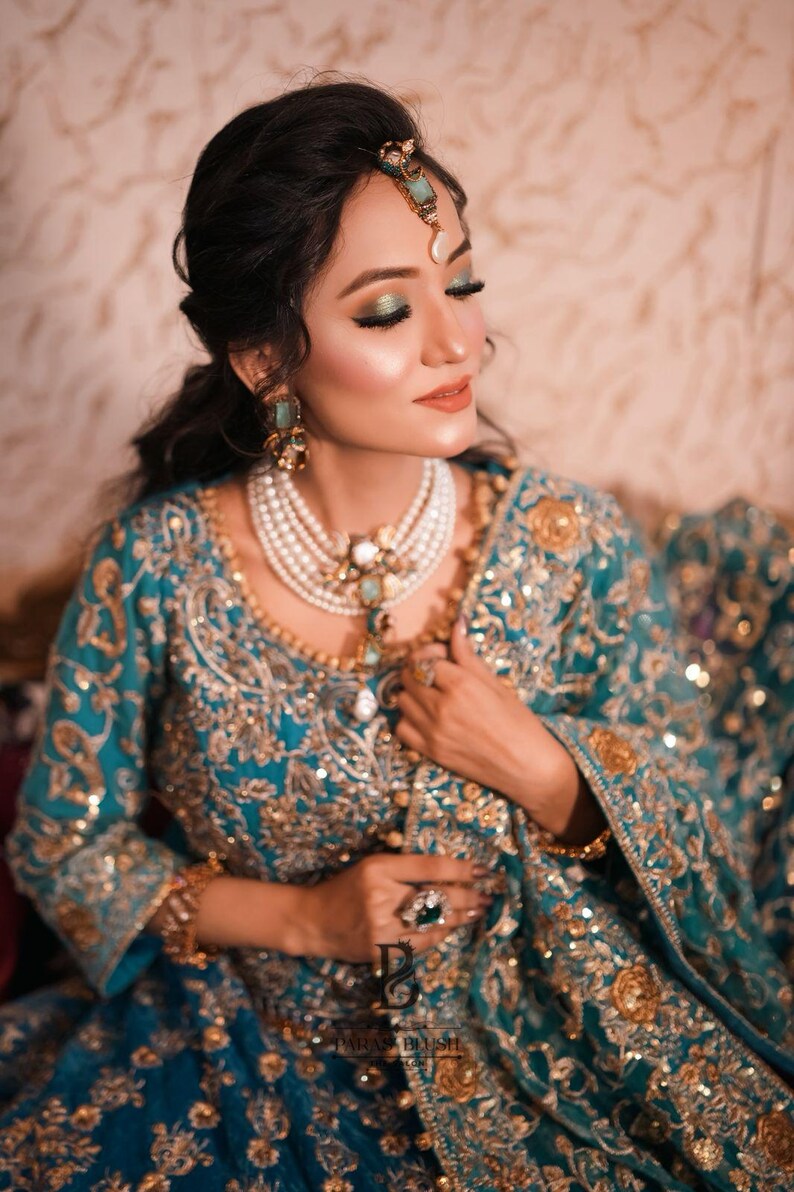 May include: A teal and gold embroidered ensemble, featuring a detailed bodice and sleeves. The outfit is adorned with gold embellishments and paired with a multi-strand pearl necklace, earrings, and a headpiece. A large ring with a green stone is visible.