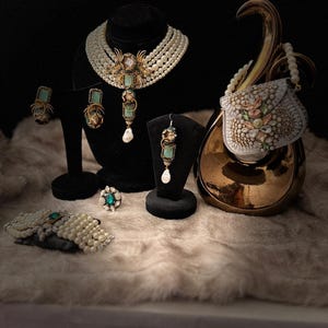 May include: An assortment of elegant jewelry pieces, including a pearl necklace, earrings, a ring, and a bracelet, displayed on a soft, textured surface. The jewelry features turquoise and gold accents, with a small, decorative purse in the background.