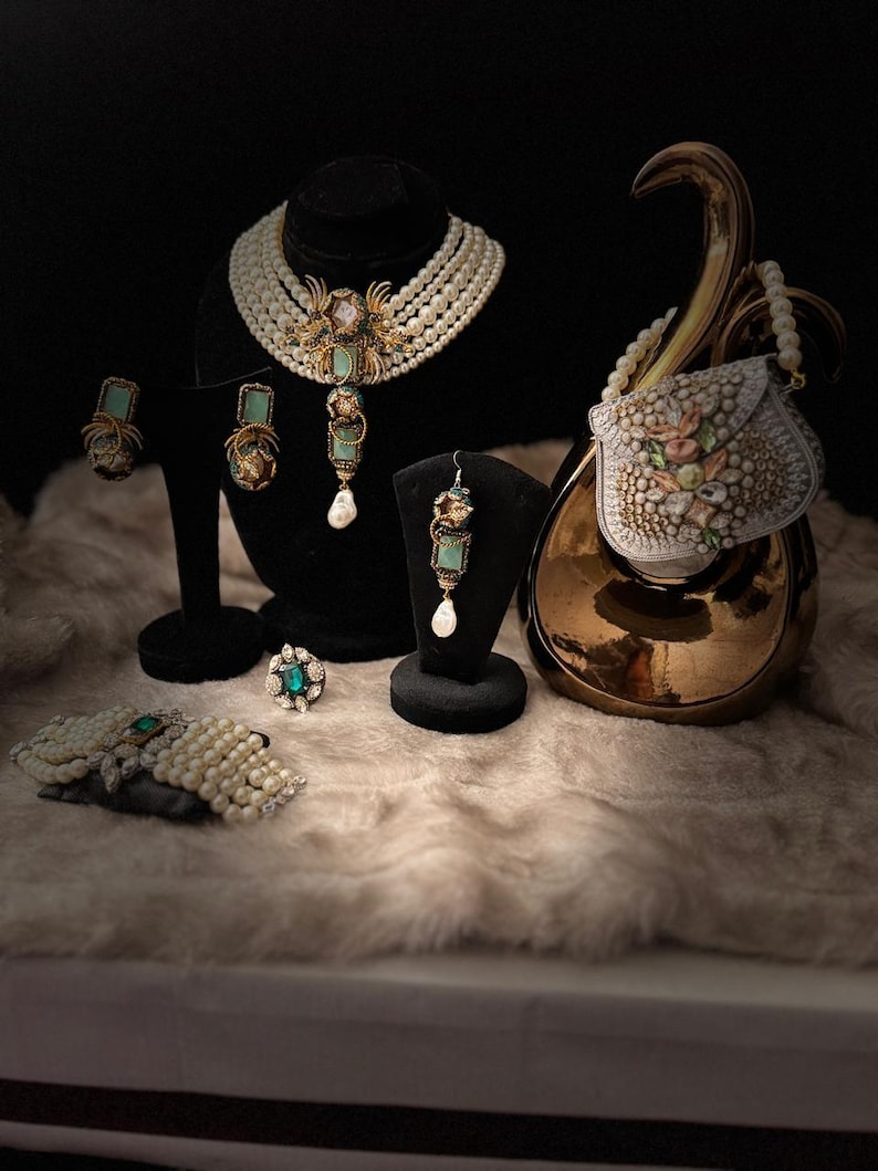 May include: An assortment of elegant jewelry pieces, including a pearl necklace, earrings, a ring, and a bracelet, displayed on a soft, textured surface. The jewelry features turquoise and gold accents, with a small, decorative purse in the background.