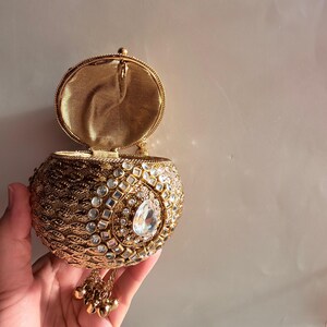 May include: A round gold-toned metal clutch bag with a textured surface, featuring a central teardrop-shaped clear crystal surrounded by smaller square and round crystals. The bag has an open lid, a gold fabric interior, and a dangling gold charm tassel.