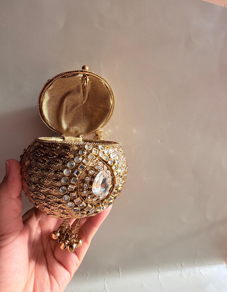 May include: A round gold-toned metal clutch bag with a textured surface, featuring a central teardrop-shaped clear crystal surrounded by smaller square and round crystals. The bag has an open lid, a gold fabric interior, and a dangling gold charm tassel.